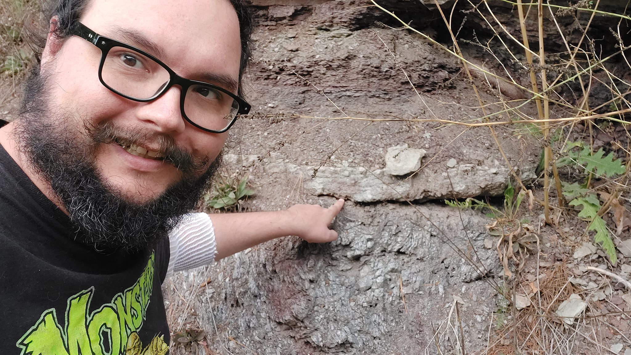 A man with short hair and bushy facial hair stands in front of a a rock outcrop on the side of the road, he is pointing to a horizontal gap that has weathered into the rock. The lower rock is grey and has no visible layers and is metamorphic, the upper rock has horizontal layering and is sedimentary